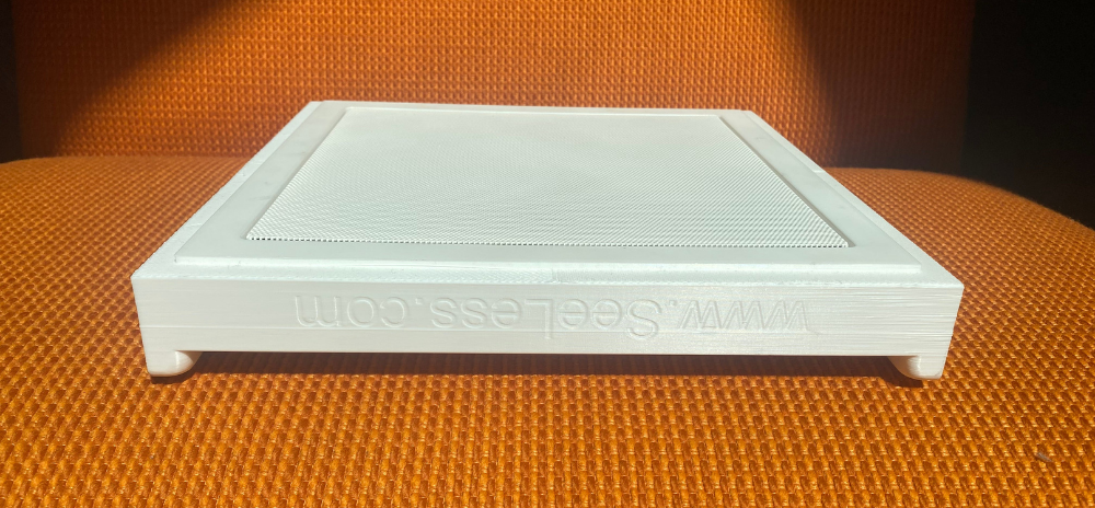 Wisdom speaker grille prototype by SeeLess, shown in a white flush-mount frame with embossed logo, placed on a textured orange surface under natural light.