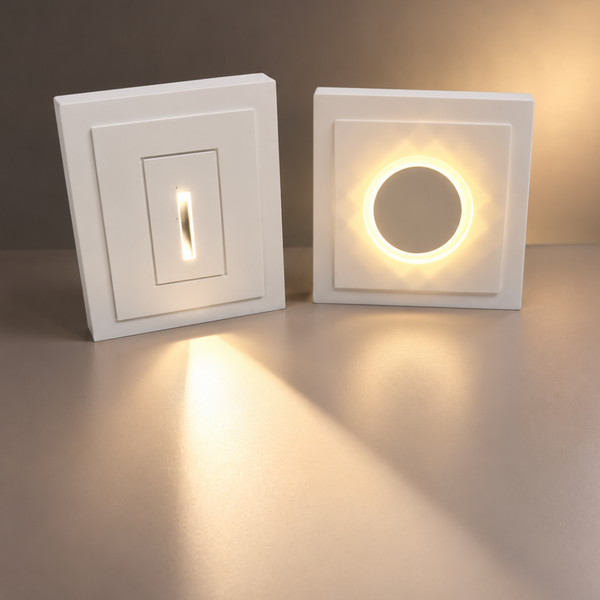 Two modern miniature white rectangular and round step/wall lights shown side by side, one with a single vertical switch and one with a circular switch, both illuminated with warm light on a neutral studio background.